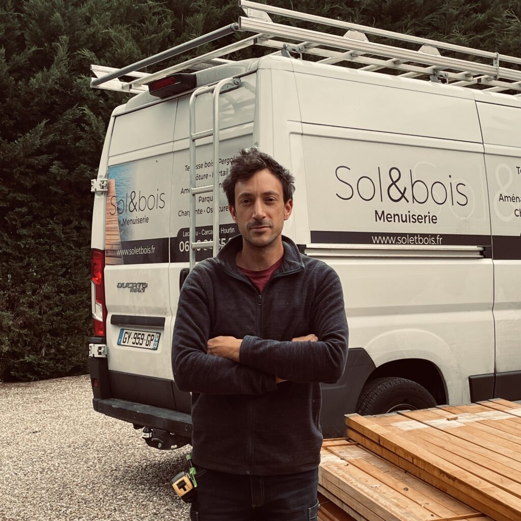 Artisan menuisier de Sol & Bois devant le véhicule de l'entreprise, à côté d'un lot de lames de bois